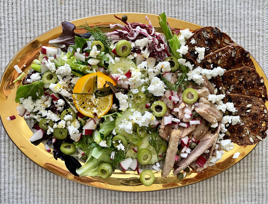 Overhead view of a summer salad featuring Mackerel - tinned fish. The salad is on a gold-rimmed plate (made by Annie Glass in Watsonville, CA) with salad greens, Castelvetrano, crackers, feta cheese, cucumbers, radishes, radicchio, Meyer lemon for dressing, fresh dill.