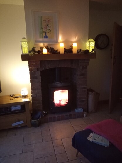 Log burner contains a roaring fire.  Fireplace built of ornamental brick with green oak mantle. On the mantle are five led candles, two in fancy candle lamps.
On the chimney breast above hangs a watercolour painting of 'Star Gazing Lilies' in a blue glass vase.