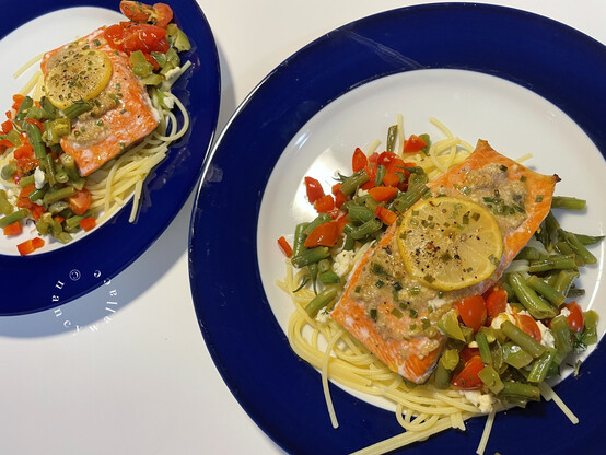 Two blue-rimmed dinner plates, each with a serving of salmon over gluten-free pasta. A lemon slide and walnut paste with chives covers the top of the salmon, and there are minced veggies as described to cover the pasta.