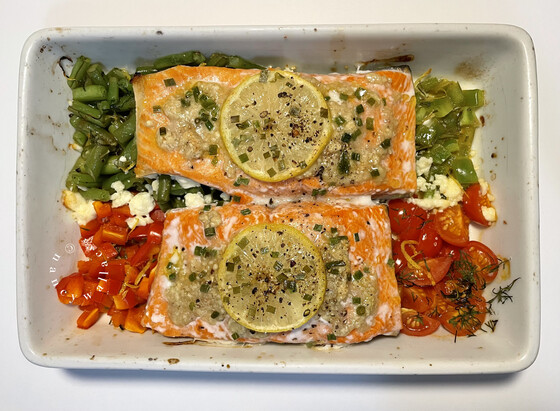 Fresh out of the oven in a white rectangle baking dish: 2 pieces of salmon covered with lemon and chives, sitting on assorted chopped veggies which include French green beans, diced green and red bell peppers, chopped cherry tomatoes and feta cheese.