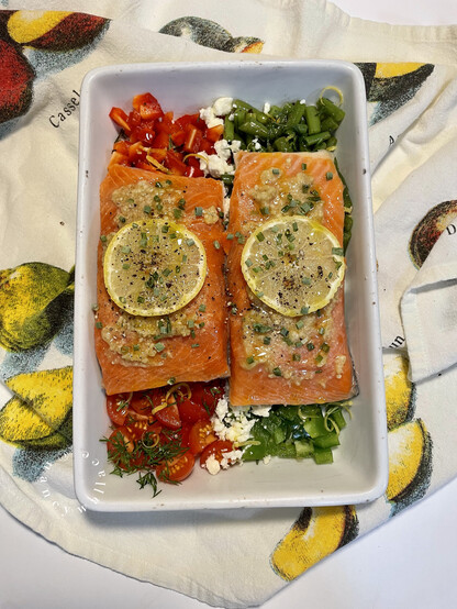 Before baking: two pieces of sockeye salmon covered with walnut paste and a lemon slice in a white rectangular baking dish, sitting on assorted chopped veggies.
