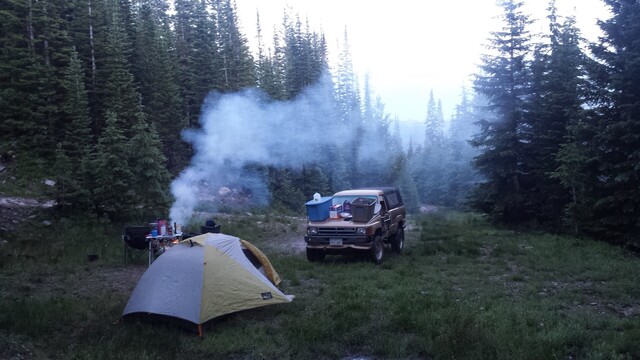 A camp setup in a meadow surrounded by conifers. It is just after sunset. There is a tent setup in the foreground. A campfire is putting up smoke. A first generation 4runner sits in the middle of the image.