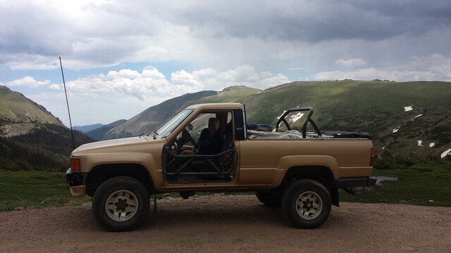A first generation 4runner with gold colored paint sits on a dirt road. The top is off the back of the 4runner. The doors have been removed from the cab. Big mountains are in the background.