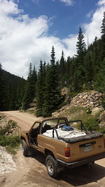 A dirt road in rocky mountain national park goes through conifers. A gold colored first generation 4runner with the top removed and the doors removed sits in the foreground with the front end pointed away from the camera.