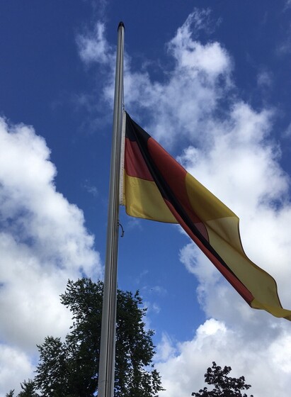 Die deutsche Flagge auf Halbmast, Im Hintergrund ein Baum und blauer Himmel mit Wolken.