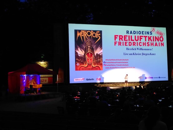 Bühne mit großer Leinwand, darauf ein Filmplakat von "Metropolis" mit der Maschine und dem Babel"-Turm.
Davor eine Person mit Mikrofon. Links daneben unter einem Pavillon ein Klavier mit Pianist.