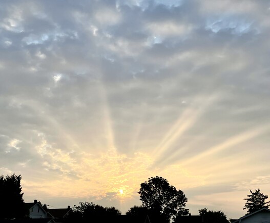 The sun just above a horizon of silhouetted roofs and trees. The sky is mostly clouded with blue breaks. Bright crepuscular rays burst from the sun in every direction and light up the clouds