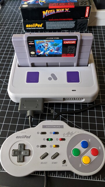 Photo of an Analogue Super Nt console with a Mega Man X cartridge sitting in it and an asciiPad controller plugged in to it. Behind the console are the boxes for Mega Man X and the asciiPad. They're all sitting on a black and white cutting mat on a desk.