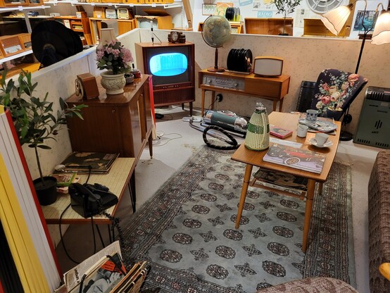 Ein altes Wohnzimmer aus den 1950ern ist nachgestellt mit Holzfernseher, Mobiliar, Geschirrutensilien, Staubsauger, Teppich.
