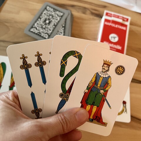Linke Hand mit 3 Scopa-Karten. Im Hintergrund sieht man den Nachziehstapel und die Kartenschachtel. 