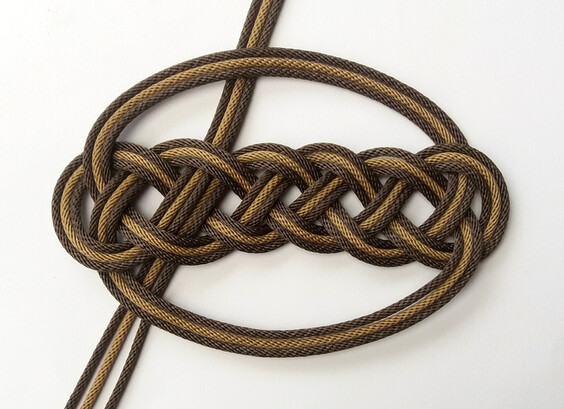 Une baderne de forme particulière.
Elle est de forme ovale, composée de :
- une petite ganse à chaque extrémité,
- une longue ganse unique sur chaque longueur.
Cette longue ganse contient quatre peties ganses, au centre, dans lesquelles les fils se chevauchent dessus-dessous.
Le nœud est triplé : un premier passage est réalisé avec de la cordelette marron foncé, un second (vers l'intérieur) avec de la cordelette de couleur beige, un troisième (toujours vers l'intérieur) est réalisé de nouveau avec de la cordelette marron foncé.