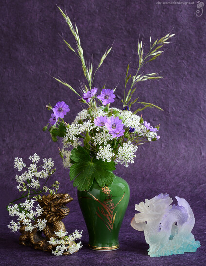 Eine grüne Vase mit violetten und weißen Wiesenblumen steht vor einem lila Hintergrund. Links von ihr versteckt sich ein bronzener Drache in weißen Blüten und niest. Rechts steht ein violetten Drache, der eine Glücksspiele in der Tatze hält.