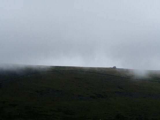 A Dartmoor ridge in heavy shadow as rains lift. The partially obscured outline of a granite Tor can be seen emerging from the low cloud and mist.