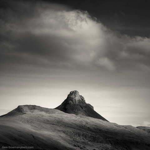 In this monochrome photograph of the Scottish Highlands, a serene scene unfolds as clouds gently gather above the iconic Stac Pollaidh. The minimalist style captures the essence of the landscape, showcasing the warm-toned tip of the mountain rising proudly against the sky. As the cloud begins to form above its peak, a sense of tranquility and grandeur envelops the frame, inviting contemplation and appreciation for the majestic beauty of nature.