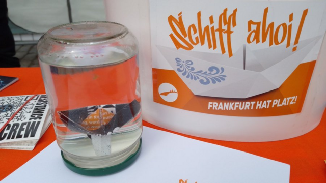 In einem umgedrehten mit Wasser gefülltem Glas ein kleines Papierfaltboot. Daneben ein großer Behälter mit einem SCHIFF AHOI! - Frankfurt hat Platz! Aufkleber. Links ragt ein Iuventa Crew Sticker ins Bild.