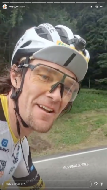 A video selfie of Strasser, smiling and wearing a white bike helmet over a white bike cap, the peak flipped up showing number 1 (he won last year). He's wearing wired earbuds and a white, yellow and black jersey as he cycles up hill.