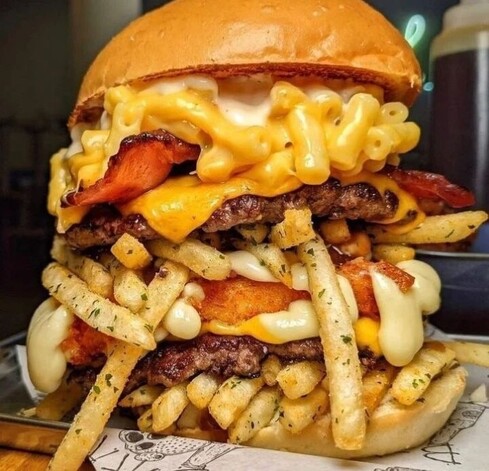 A double layered cheeseburger in a toasted brioche bun, loaded with mac & cheese, whole bacon rashers, something that looks a slice of fried chicken, fries and an unspecified type of cheese (possibly gruyère) 🧀 😋