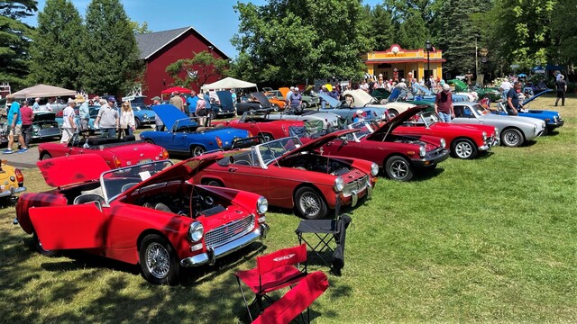 A line of brightly colored MG Midgets in the sun on a grassy lawn with a barn-like and a vintage gas station structure behind.