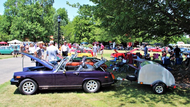 A purple rubber-bumper MG Midget on a lawn with a silver teardrop trailer behind. The Midget is to the left of the image and pointed left towards a paved pathway. The teardrop trailer is hooked up to the rear of the midget and in the shade under a tree.