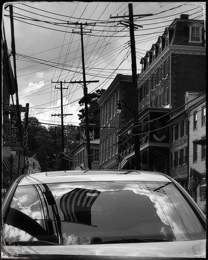Black and white photo of a car windshield with clouds, sky, and the American flag reflected on it. Above are several utility poles with a jumble of wires in front of a cloudy sky. Old timey building recede to the background on the right