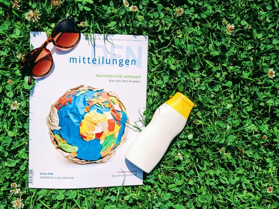 Das Foto zeigt das Magazin des DFN-Vereins „DFN-Mitteilungen“, Ausgabe 103. Das Titelbild zeigt eine Papierweltkugel. Die Teaser auf dem Titel lauten: Konnektivität weltweit – Start der GN-5-Projekte; Sicher FAIR – IAM4NFDI in der DFN-AAI; Lebenslang gültig – Das Konzept edu-ID. Das Heft liegt auf einer Wiese. Links oben auf dem Heft liegt eine braune Sonnenbrille, rechts unten eine weiße Plastikflasche mit Sonnenmilch.