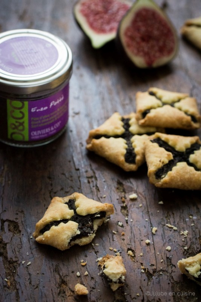 en gros plan sur une table en bois foncé, un pot de tapenade aux olives noires et aux figues, des figues fraîches coupées en deux, et à l'avant plan des jolis biscuits fourrés. la pâte est repliée, chaque coin vers le centre en laissant un espace, du coup ça fait un motif de croix laisse qui voir la tapenade foncée dedans. 

le tout a l'air gourmand, avec une texture de sablé et on devine que ça sera goûtu et salé. 
