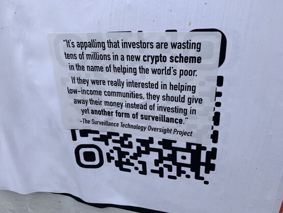 A label that has been slapped over a big QR code. The label has a quote by Albert Fox Cahn, the executive director of the Surveillance Technology Oversight Project, that says “It’s appalling that investors are wasting tens of millions in a new crypto scheme in the name of helping the world’s poor,. If they were really interested in helping low-income communities, they should give away their money instead of investing in yet another form of surveillance.”