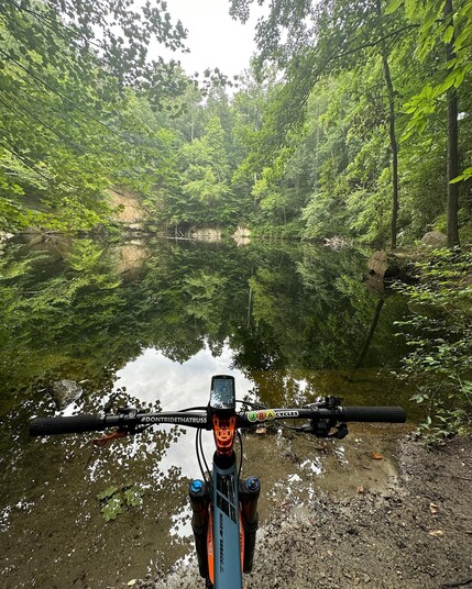 Mountain bike from the rider’s perspective in front of a quarry pond with lush woodlands surrounding it. There are two stickers in the bike’s handlebars. The left one reads #DontRideThatRuss. The right one says JRA Cycles.