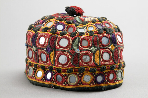 Hat; small domed hat intensely decorated with mirror work and Image of item 1317 multicoloured buttonhole stitched motifs outlined in black on a red ground. The top of the hat is finished with a small black and red tassel.