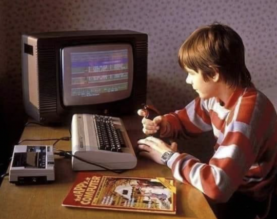 Teenager am Schreibtisch mit Commodore C64, Fernseher, Datasette und Happy Computer Magazin. Er selbst hat einen Joystick in der Hand