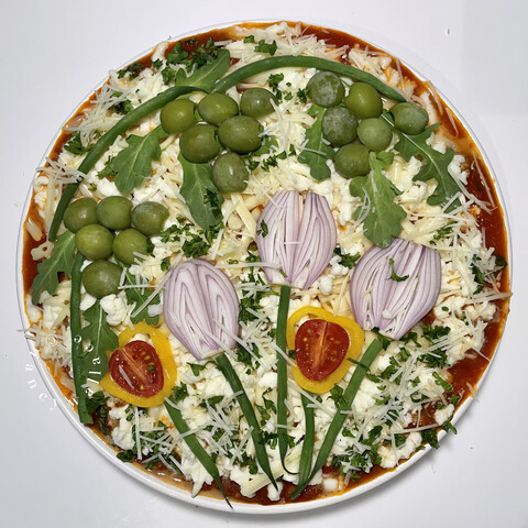 Overhead view of an unbaked lasagne that has been decorated with green olives to look like grape vines. Also with red onions and tomatoes as flowers.
