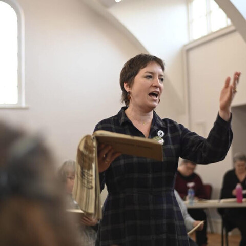 Caro, a woman with short hair, is leading a shapenote song: holding the songbook in her right hand, her left hand held up, beating the time of the song. In the foreground and background you can see other shapenote singers sitting in the hollow square, the typical arrangement for a shapenote singing. 