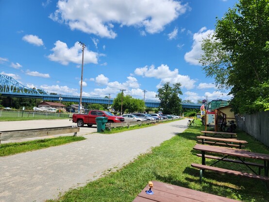 Blue sky with small clouds above a parking lot with a few cars beside a gravel bike trail and some picnic tables.
