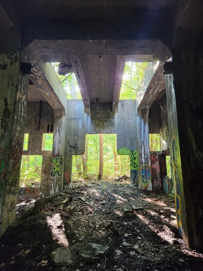 View from inside deteriorated heavy concrete structure. Large openings reveal glowing green woodlands past the walls. Various spray paint art, mostly in blue and neon green cover much of the interior.