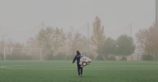 Mensch mit Trainingsanzug und hellen Turnschuhen trägt ein Netz mit Bällen über einen Fußballplatz. Das Wetter: diesig.