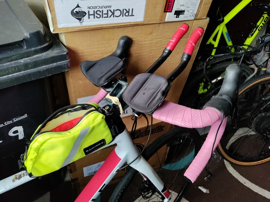Close up photo of a road bike with clip on TT bars attached. The photo is taken in a garage, surrounded by other bikes, cardboard boxes and a wheelie bin. 