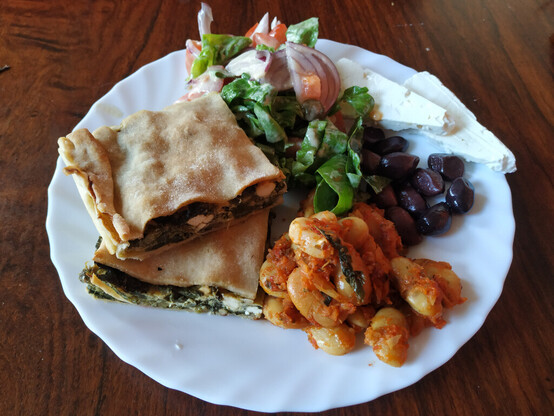 A white plate with two slices of a hortopita (green leaf and feta cheese pie), green salad, olives, feta and baked broad beans.
