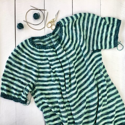 A flat lay view of a knit striped tee in teal and light turquoise yarn. It's pictured on a white plank background, together with 2 tiny circular needles, 2 balls of dark yarn, and scissors.