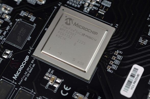 An angled macro photograph of a Microchip PolarFire SoC system-on-chip, installed in a black circuit board labelled as the PolarFire SoC SEV Kit but better known as the PolarFire SoC Video Kit. Teh chip is beneath an aluminium heatspreader, labelled with the Microchip logo, various code numbers, and the RISC-V logo.