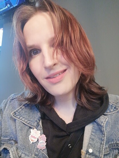 Selfie von Lili vor einer grauen Wand. Sie lächelt leicht. Das Haar liegt ungekämmt halb im Gesicht, oben zum Mittelscheitel mit leicht Volumen im Pony. Man sieht nur etwas grauen Lidschatten auf den Augen, sonst komplett ungeschminkt.

Sie trägt eine Jeansjacke und darunter einen schwarzen Hoodie.