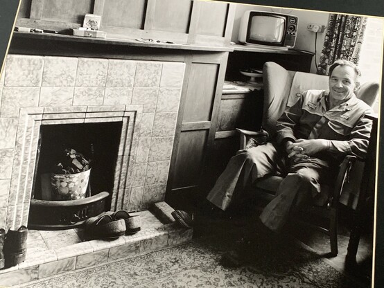 A photograph of a man sat in his living room in black and white, called “Mr. Clements At Home”. There is a retro TV in the background.