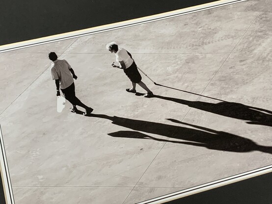 Two figures walking across an open area, casting long shadows, in black and white. This one is called “Shadows in the Evening”.