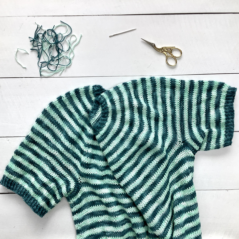 A flat lay view of a knit striped tee in teal and light turquoise yarn. It's pictured on a white plank background, together with lots of snipped of yarn, a sewing needle, and scissors.