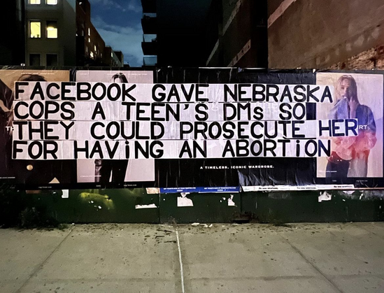 street pasteup of black text on white background that read: FACEBOOK GAVE NEBRASKA COPS A TEEN'S DMS SO THEY COULD PROSECUTE HER FOR HAVING AN ABORTION

The pasteup is on a fence against a sidewalk, pasted over a four or five other poster ads. In the background there are a series of urban buildings and a small bit of dusky sky. 