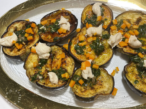 Roasted eggplant slices on a clear glass plate with a gold rim. The eggplant is topped with oregano, chopped yellow bell peppers and drops of tahini. The plate is made by Annie Glass, Watsonville, CA. 
