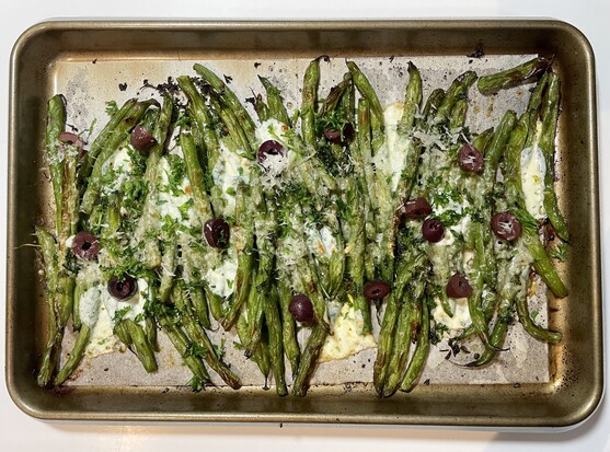 A sheet pan of roasted green beans fresh out of the oven, dotted with kalamata olives, parsley and melted parmesan cheese and mozzarella pearls. 
