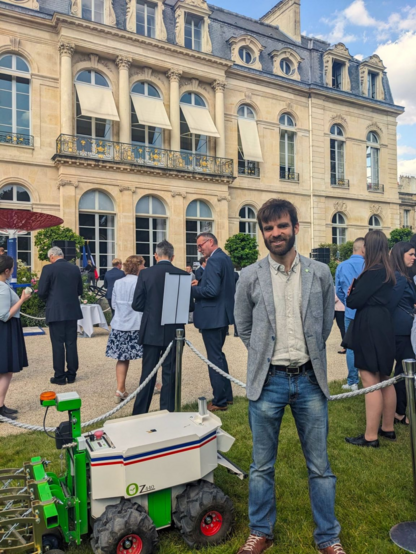 Gaëtan Severac, cofondateur de Naïo Technologies, debout. Il se trouve devant le bâtiment de l’Élisée, à coté du robot Oz.