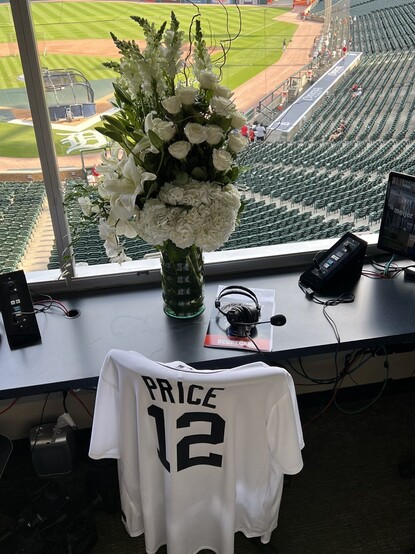 A Jim Price Jersey is hanging on the back of his chair in the Comerica Park press box. A gorgeous bouquet of white roses and other flowers (carnations and glads I think) is in front of the chair, and his headphones are on the desk. The field is visible through the window.