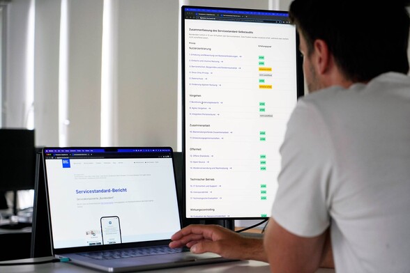 A person standing at a desk in a modern office space, looking at two screens – one says: ‘Service standard report’ in German, the other shows a long list of things that are colour-coded with different colours
