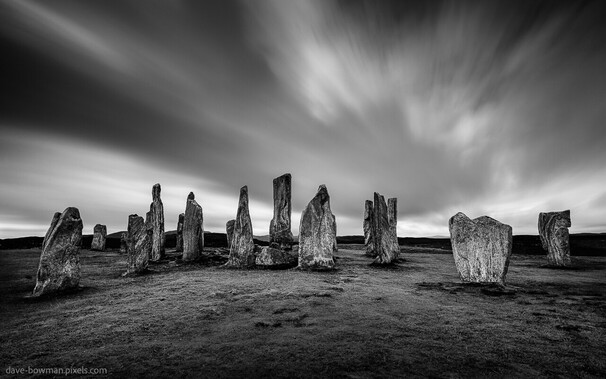 A black and white photograph of the majestic Callanish Standing Stones on an overcast day on the Isle of Lewis, Outer Hebrides of Scotland. The stones, weathered by centuries of existence, stand resolute against the backdrop of a grayscale canvas, their solemn grandeur accentuated by the long exposure technique that blurs the passage of time. Wispy clouds gracefully meander over the scene, casting a gentle shadow and infusing an air of melancholy, as if the very landscape echoes with the ancient whispers of history and mystery.